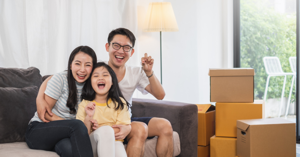 Asain family smiling holding keys to new house with packing boxes around them.  